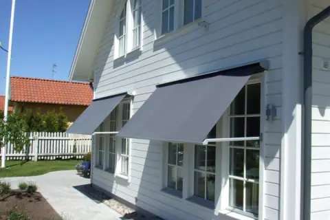 Grå markiser monterade på ett vitt trähus ger skugga åt fönstren. En vacker trädgård med stenläggning och staket syns i bakgrunden.
