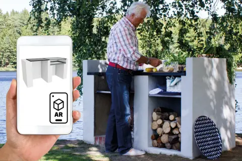Man förbereder mat vid en murad grillplats vid en sjö. En mobil visar en 3D-modell av grillplatsen i AR.