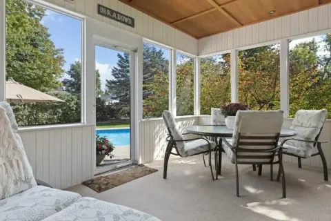 En inglasad altan med utsikt över en pool och trädgård. Ett runt bord med stolar står redo för fika.