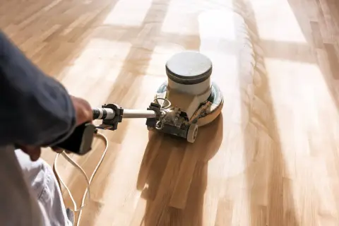 En person slipar parkettgolv med en maskin. Dammet virvlar upp när golvet blir rent och redo för behandling.