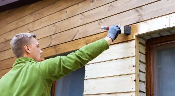 Man målar panel utomhus med en pensel. Träet är ljust och fräscht.