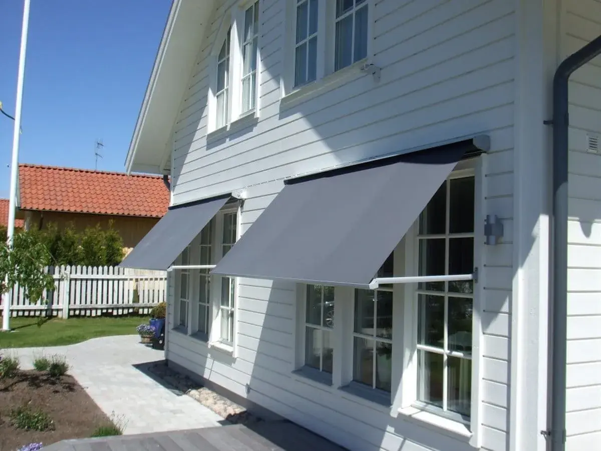 Grå markiser monterade på ett vitt trähus ger skugga åt fönstren. En vacker trädgård med stenläggning och staket syns i bakgrunden.