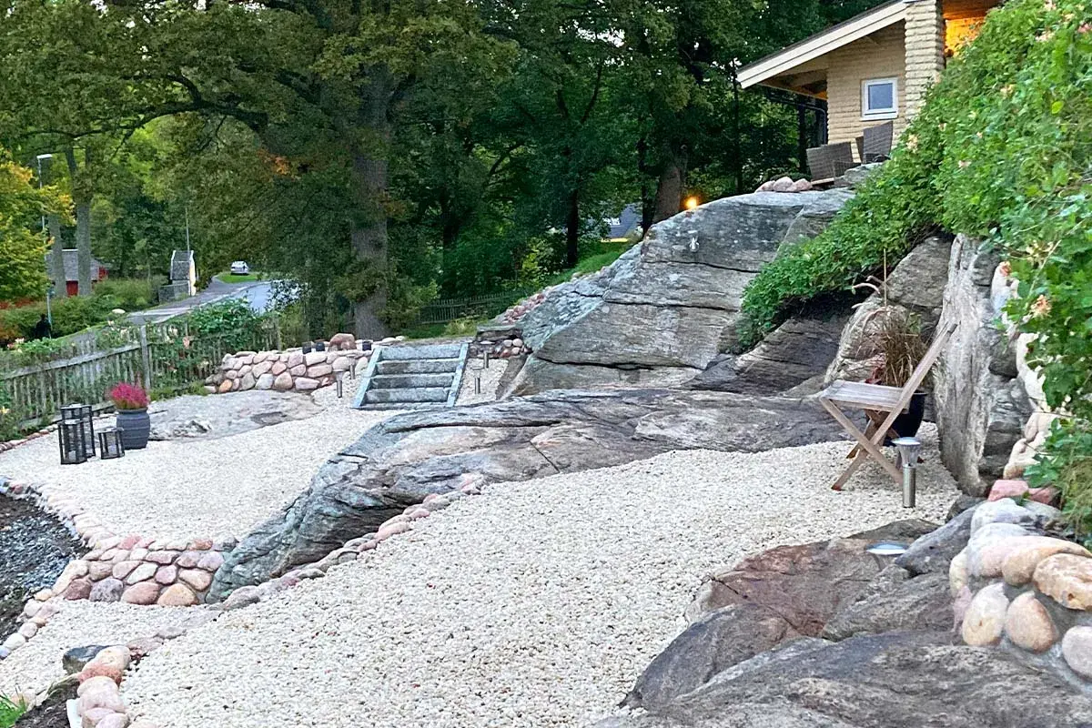 En vacker stenlagd uteplats med grusgångar, stenmurar och trappor som leder upp mot ett hus. En inspiration för en naturlig och robust utomhusmiljö.