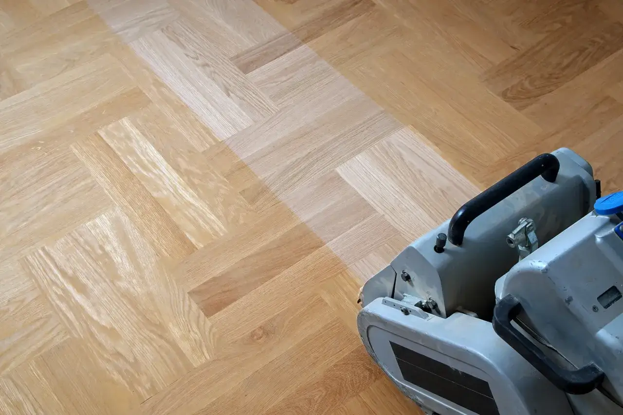 Maskin slipar parkettgolv i fiskbensmönster. En del av golvet är ljusare där slipningen pågår.