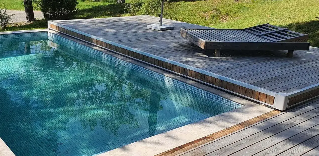 En pool med bl&aring; mosaik och en tr&auml;altan. En solstol st&aring;r p&aring; altanen, perfekt f&ouml;r att njuta av solen efter att ha byggt eget poolskydd.