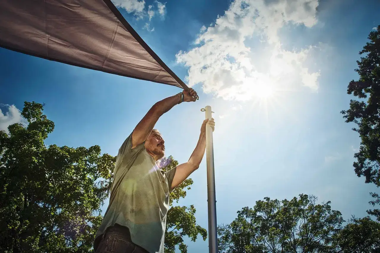 Man s&auml;tter upp ett solsegel mot en klarbl&aring; himmel med solens str&aring;lar som lyser. Tr&auml;d syns i bakgrunden.